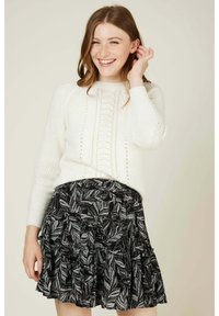 Pull en tricot blanc avec un motif texturé, associé à une jupe noire avec un imprimé de feuilles argentées et des volants superposés.