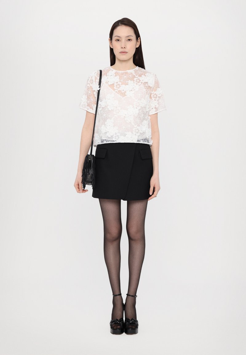 Witte bloemenkant top, zwarte wikkelrok, zwarte panty en plateau schoenen. De outfit bevat texturen en contrasterende kleuren.