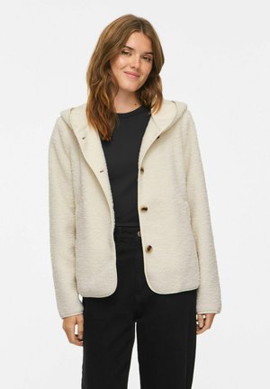 Giovane donna che indossa una giacca in pile color crema con bottoni e cappuccio sopra una camicia nera e pantaloni neri, in piedi davanti a uno sfondo bianco.