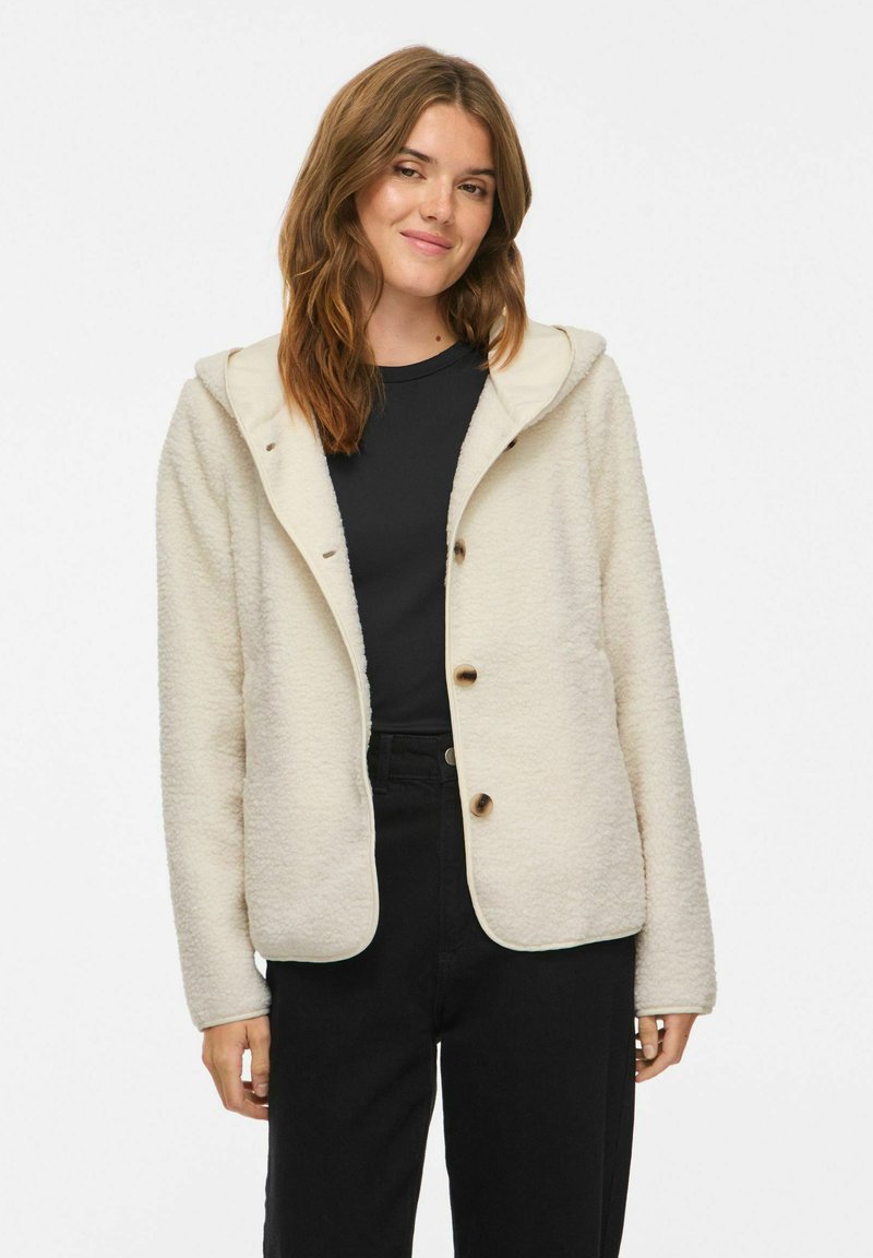 Jeune femme portant une veste polaire crème boutonnée avec capuche par-dessus une chemise noire et un pantalon noir, debout devant un fond blanc.