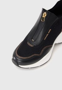 Baskets noires à enfiler avec fermeture éclair dorée, dessus en mesh, accents en cuir, bordure marron, semelle blanche et logo "Michael Kors" sur le côté.