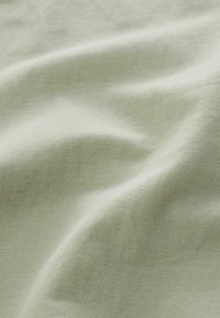 Šviesiai žalia tekstūruota medžiaga su matoma smulkia pynimo struktūra ir minkštais klostėmis, kurios sukuria švelnias šešėlius.