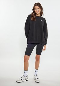 Felpa nera oversize con logo ricamato, abbinata a pantaloni bici neri. Sneakers bianche con dettagli neri e calze a righe.