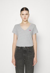 Szürke V nyakú pamut póló, rövid ujjakkal, a mellkason egy kis piros Levi's logóval, magas derekú fekete farmerral párosítva.