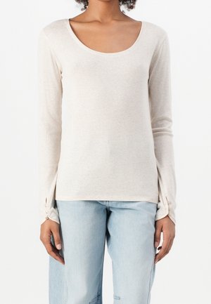 Vrouw draagt een beige lange mouwen top met een ronde hals en knoopdetails op de manchetten, gecombineerd met lichtblauwe high-waisted jeans, staand tegen een witte achtergrond.