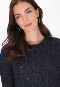 Laivansiniset neulepusero pyöreällä kaula-aukolla, jossa on teksturoitua kangasta ja hajallaan pieniä paljeteja hienovaraisena yksityiskohtana.