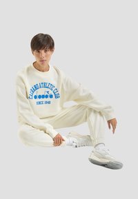 Completo felpa e pantaloni della tuta color crema con grafica blu "Cabrano Athletic Club", abbinato a sneaker bianche. Texture liscia, vestibilità comoda.