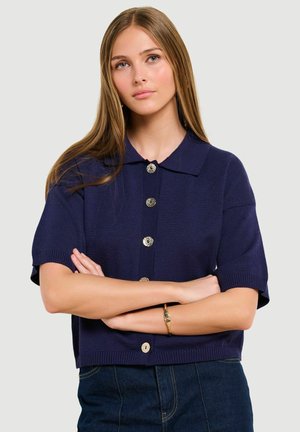 Mladá žena s dlhými svetlohnedými vlasmi, oblečená v námornícky modrom krátkym svetríku na gombíky a tmavých džínsoch, so založenými rukami a neutrálnym výrazom.