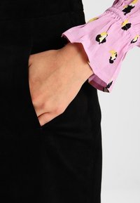 Camisa de manga larga de tela rosa con un patrón negro y amarillo. La camisa tiene un fruncido en la muñeca y se lleva con material negro.