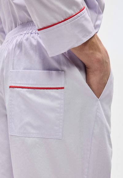 Αρσενικό χέρι στην τσέπη των ανοιχτόμωβ παντελονιών με κόκκινο περιθώριο στην πίσω τσέπη και μανίκι πουκάμισου σε τόνους που ταιριάζουν με κόκκινη επένδυση.