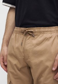 Homme portant un pantalon beige à cordon et un t-shirt noir, avec les mains visibles reposant sur les côtés.