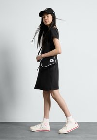 Abito t-shirt nero, sneakers bianche con accenti rossi, cappellino nero e borsa a tracolla piccola con un logo rotondo, caratterizzata da un design semplice.