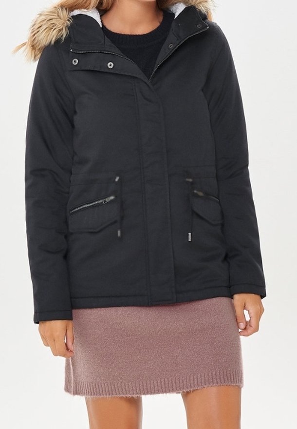 Giacca nera isolata con cappuccio in pelliccia sintetica rimovibile, tasche con zip e a toppa, e una texture liscia, abbinata a una gonna lavorata a maglia rosa.