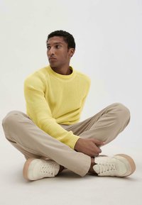Gelber Strickpullover mit Rundhalsausschnitt und geripptem Muster, kombiniert mit hellbeigen Hosen und cremefarbenen Sneakers mit Plattformsohle.