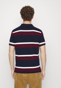 Námořnická modrá polo košile s horizontálními vínovými a bílými pruhy. Má límec a krátké rukávy. Hladká tkanina s přiléhavým střihem.