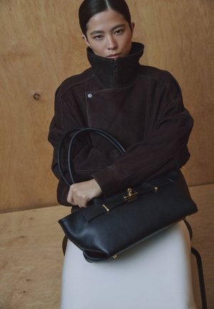 Zwarte leren handtas met gouden beslag, gladde textuur en een gestructureerd ontwerp, gedragen door een persoon die een bruine suède jas draagt.