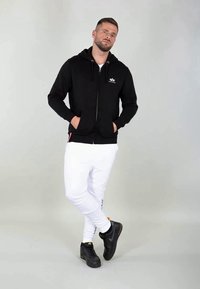 Man wearing black zip-up hoodie and white pants, standing with hands in pockets and one leg crossed over the other against a gray background.
