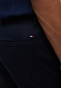 Gros plan d'une personne portant un pantalon bleu marine avec une poche arrière boutonnée et un petit logo rouge, blanc et bleu sur la poche.