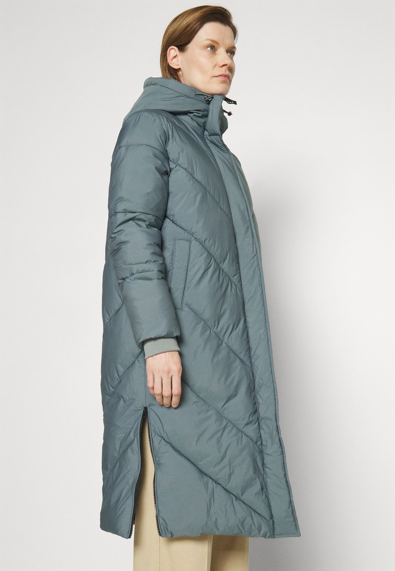 Abrigo largo acolchado tipo puffer en teal claro con un alto cuello, cremalleras laterales y patrones de costura diagonales, que presenta un ajuste relajado y holgado.