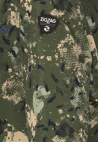 Grøn camouflagejakke med tekstureret stof, zigzag-logo-patch og en blanding af creme og marineblå accenter i et moderne mønster.
