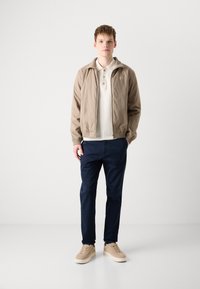 Light beige zip-up jacket, white collared shirt, navy pants, and tan sneakers. Model stands against a plain white background.