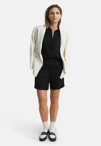 Witte streepblazer over een zwart overhemd met knopen en zwarte shorts. Gecombineerd met blauw-witte instappers en witte sokken.