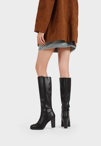 Stivali neri in pelle al ginocchio con tacco a blocco, caratterizzati da un dettaglio in fibbia argentata e una finitura liscia, abbinati a un cappotto in suede marrone.