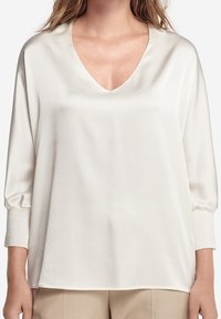 Blouse blanche à col en V en tissu léger, avec des manches trois-quarts à poignets froncés et une coupe décontractée.