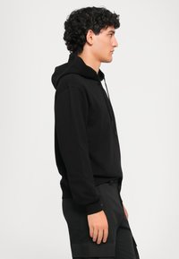 Jovem com cabelo encaracolado, vestindo um moletom preto e calças cargo pretas, virado para a direita contra um fundo claro e simples.