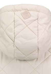 Capuche de veste matelassée beige clair avec motif losange cousu et écusson logo "Sub Level" embossé.