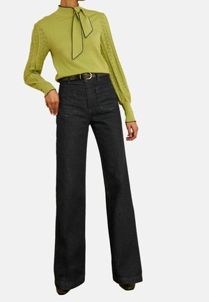 Femme portant un haut en maille vert citron à manches longues avec un col à nœud papillon, associé à un pantalon noir taille haute à jambes larges et une ceinture noire.