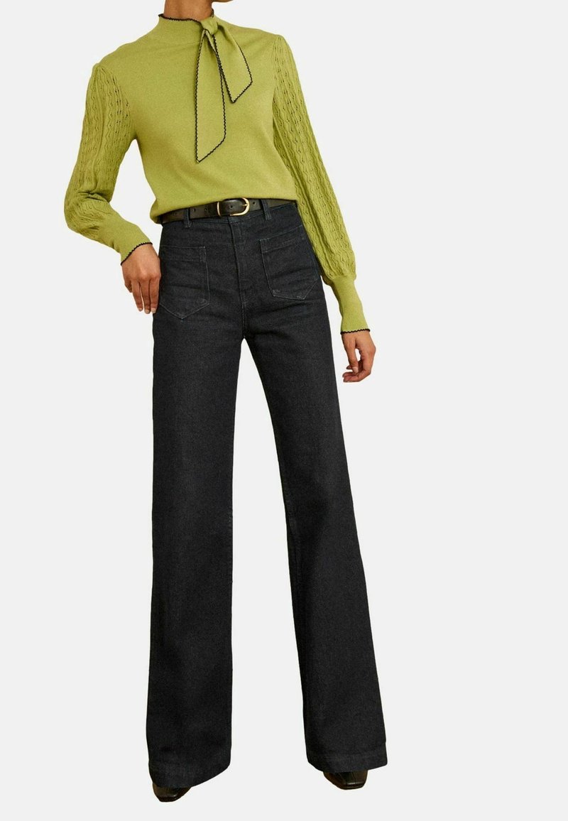 Femme portant un haut en maille vert citron à manches longues avec un col à nœud papillon, associé à un pantalon noir taille haute à jambes larges et une ceinture noire.