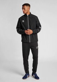 Svart sportjacka med dragkedja, med vita ränder och logotyp. Matchas med svarta byxor och blå sportskor. Slät tygstruktur.