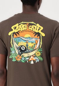Braunes Baumwoll-T-Shirt mit einem bunten Grafikmotiv eines Billardballs in einer Hängematte, tropischen Pflanzen, Wellen und kräftigem Text darüber.