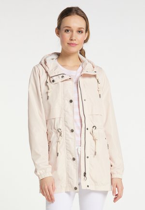 Jeune femme portant une veste à capuche beige clair avec cordons et boutons-pression, debout devant un fond blanc uni.