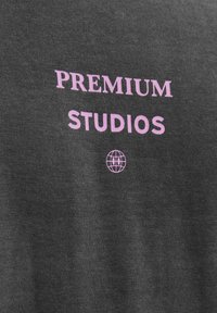 Sivi materijal sadrži riječ "PREMIUM" u roza boji iznad "STUDIOS" i malom ikonom globusa, sve otisnuto u podebljanom, modernom fontu.