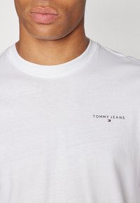 T-shirt de algodão branco com decote redondo e um pequeno logótipo bordado em preto "TOMMY JEANS" com um destaque em vermelho e azul. Textura suave.