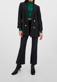 Blazer nero con design doppiopetto, bottoni bianchi e vestibilità su misura. Abbinato a un top corto a coste verde e jeans neri a zampa.