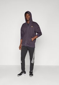 Sweatshirt roxo com fecho éclair e capuz, apresentando um logótipo laranja. Combinado com calças de moletom pretas com riscas brancas nas laterais e sapatos pretos.