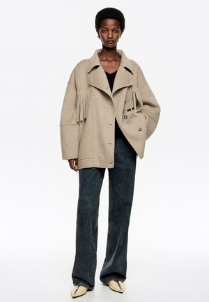 Femme portant une veste à franges beige surdimensionnée, un haut noir, un jean large foncé et des chaussures beiges pointues sur fond blanc.