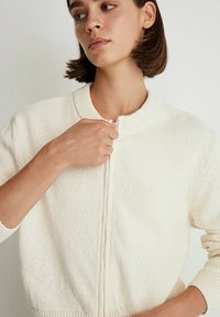Cream-colored knitted cardigan with a round collar, zipper closure, and a textured finish, featuring ribbed cuffs and hem.