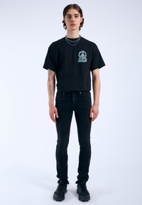 Camiseta negra con diseño gráfico turquesa en el pecho, combinada con jeans negros ajustados y zapatillas negras texturizadas. Estilo minimalista con tatuajes visibles.