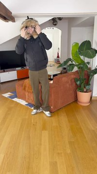 Μαύρο φούτερ μπουφάν με κουκούλα από τεχνική γούνα, χακί παντελόνι και ασημένια αθλητικά παπούτσια; στέκεται σε ξύλινο πάτωμα σε δωμάτιο με φυτά και έπιπλα.