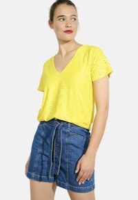 Dzeltena īsām piedurknēm V-veida kakla blūze ar acs detaļām, apvienota ar augsto jostasvietu ziliem džinsiem, kuriem ir sasietā josta un kabatas.