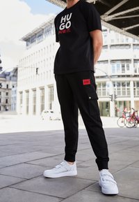 Zwart T-shirt met witte en rode tekst, gecombineerd met zwarte cargobroek met een rood logo patch. Witte sneakers met bandaccenten.