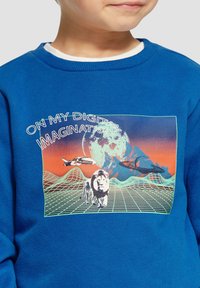 Niebieska bluza z grafiką przedstawiającą lwa, statki kosmiczne i górzyste tło. Napis brzmi "W MOJEJ CYFROWEJ WYOBRAŹNI."