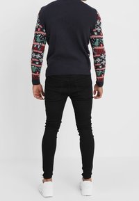Pull en tricot marine avec des manches à motifs festifs comportant des designs rouges, verts et blancs ; associé à un pantalon skinny noir et des chaussures blanches.