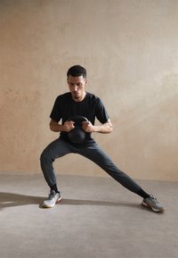 Czarna koszulka i ciemnoszare dresy noszone przez osobę wykonującą ćwiczenie z kettlebellem. Kettlebell jest czarny z wyprofilowanym uchwytem.