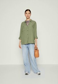 Zeleno gumbasto srajco z žepoma na prsih, svetlo modre široke hlače ter rjav teksturiran ročni torbico. Model stoji ob svetli ozadju.