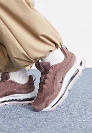 Osoba ubrana w beżowe spodnie i białe skarpetki z wrzosowymi butami Nike Air Max na niebieskim dywanie.